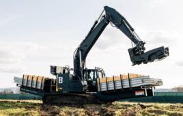 A machine resembling a backhoe has picks up a steel beam from a shelf built onto its side.