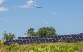 An airplane flies in a partly cloudy sky above trees and a solar panel array.