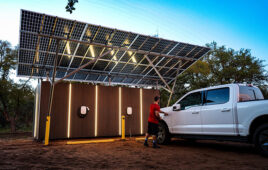 A person is plugging an electric vehicle charger into a truck that is parked beneath a canopy of solar panels.