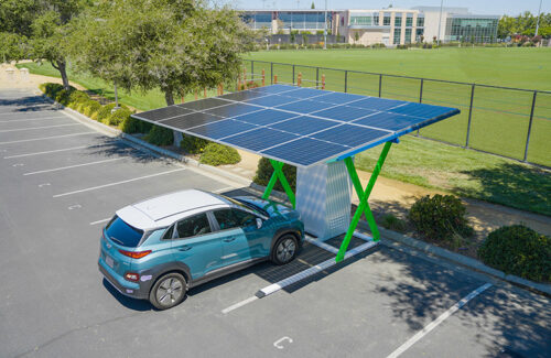 A compact car is parked under a small solar canopy and fitted with an electric car charger.
