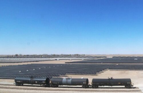 Train cars pass by a field of solar panels on a dry landscape. 